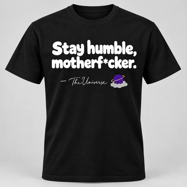 A black crewneck T-shirt mockup with “Stay humble, motherf*cker.” across the front, with a smaller attribution dash, signature and The Universe mascot beneath. Complete shirt shown against the supplied studio background.